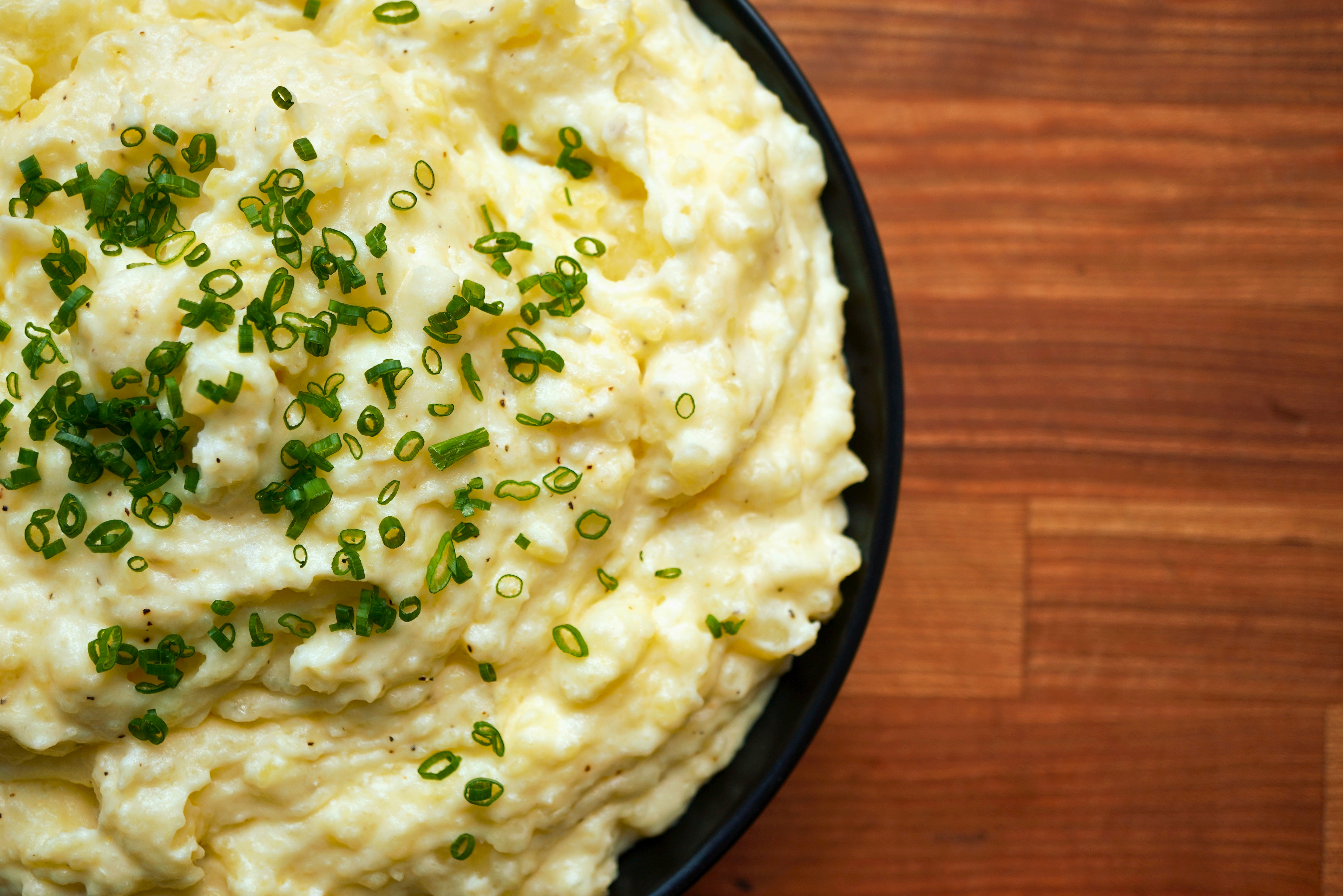 A bowl of creamy mashed potatoes garnished with chopped green onions on a wooden table.