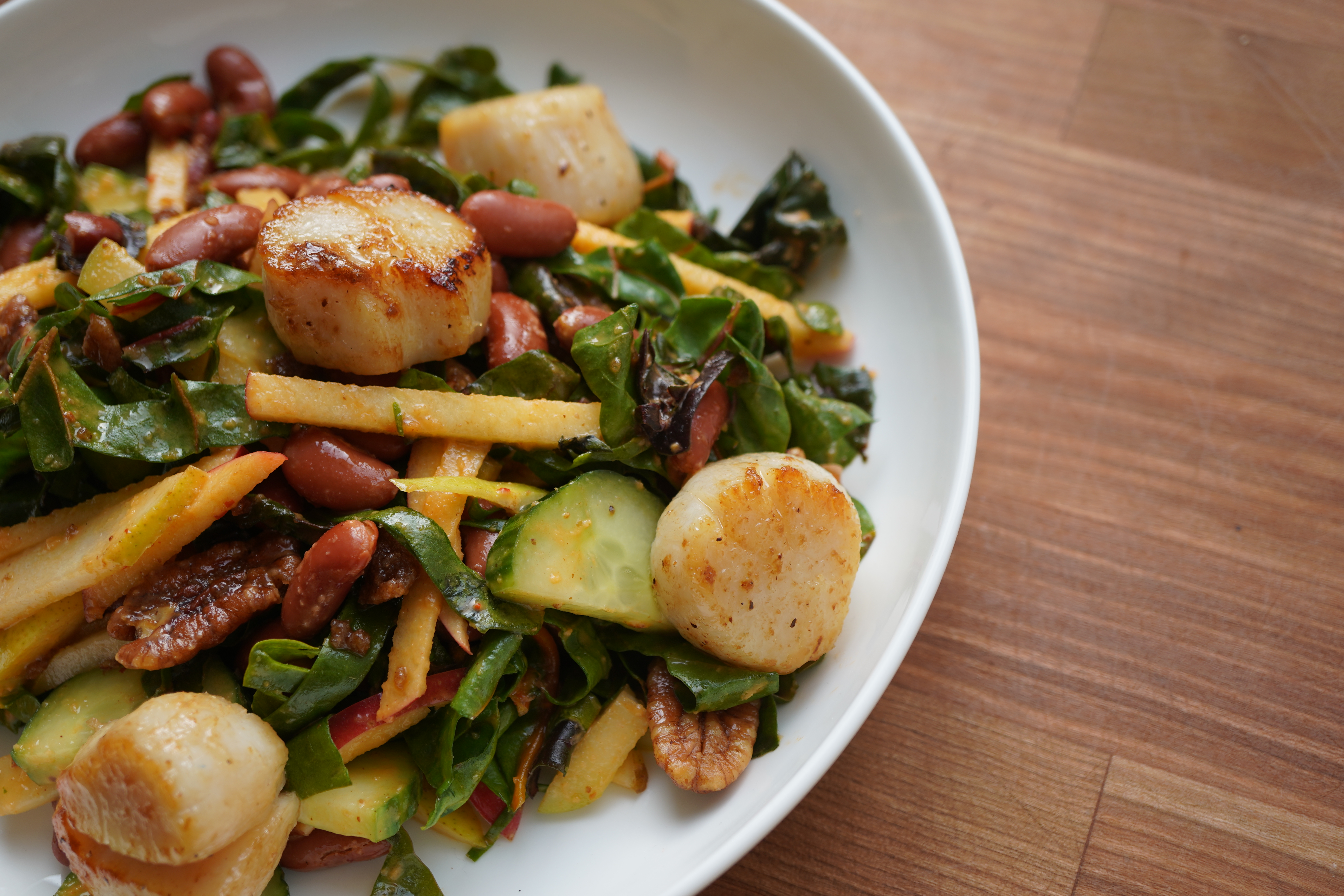 A plate of seared scallops, roasted vegetables, and greens, arranged on a wooden table.