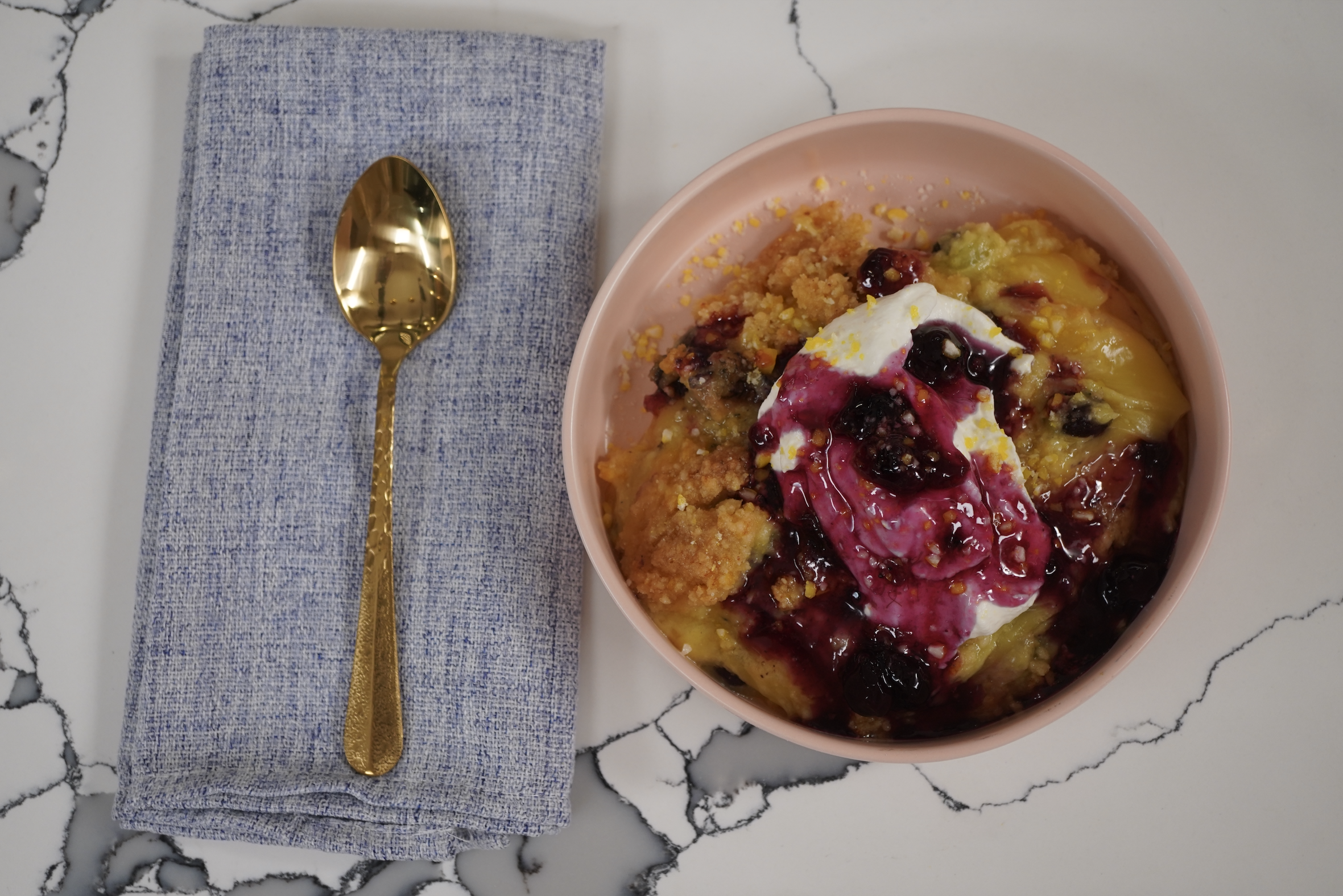 A pink bowl filled with a colorful porridge-like dish, topped with a dollop of cream or yogurt and berries, accompanied by a gold spoon on a gray cloth napkin.