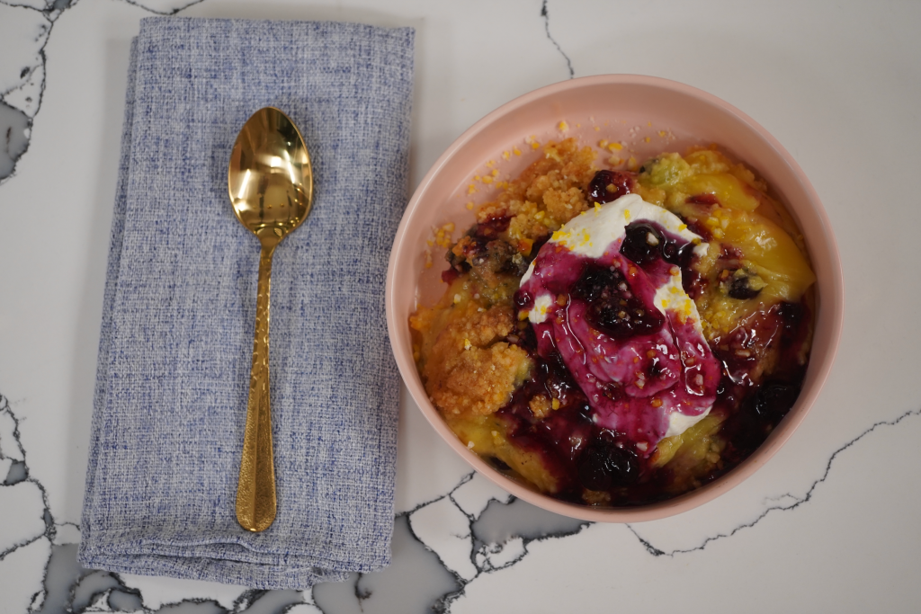 A pink bowl filled with a colorful porridge-like dish, topped with a dollop of cream or yogurt and berries, accompanied by a gold spoon on a gray cloth napkin.