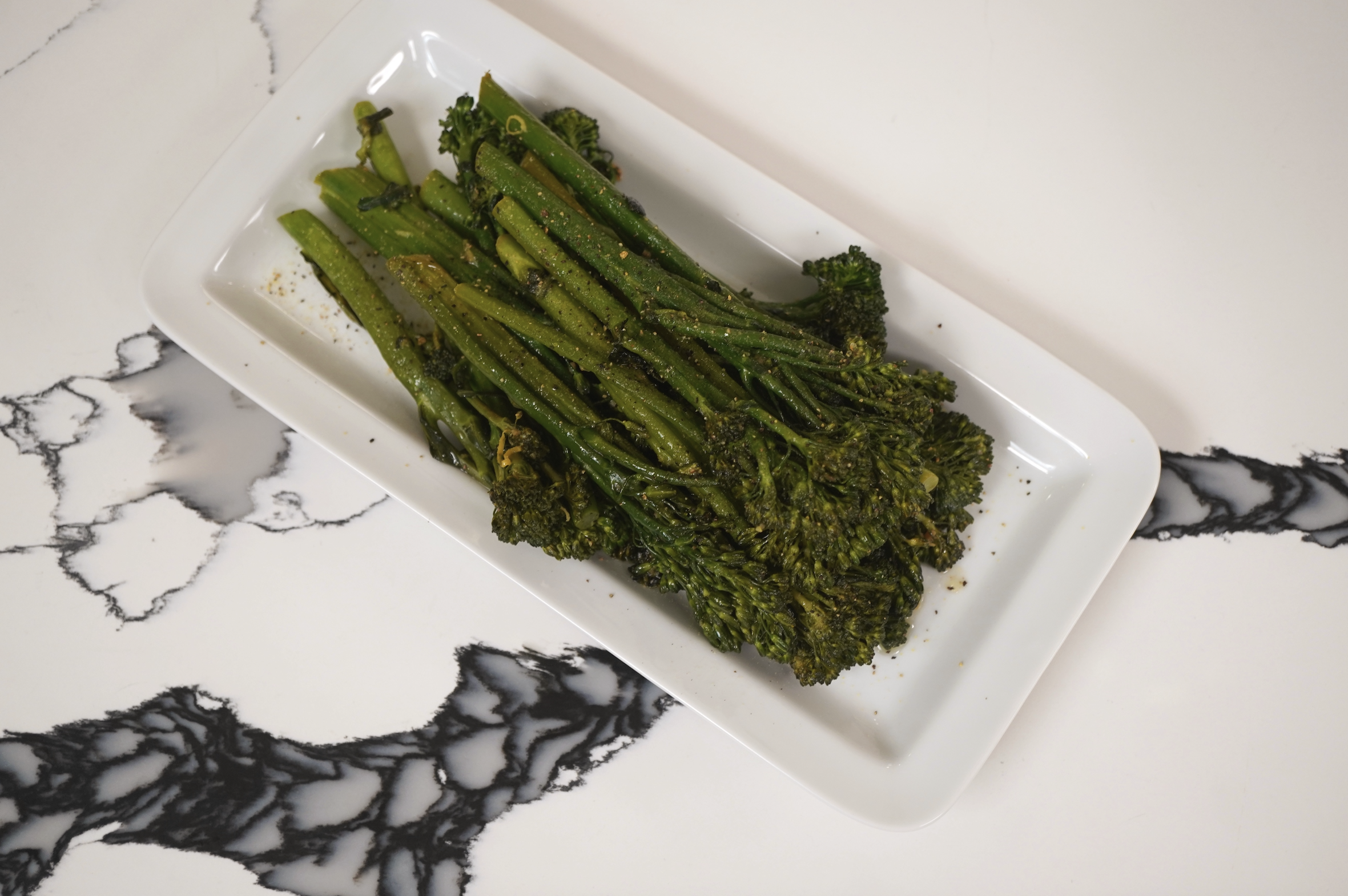 Plate of roasted green vegetables, including asparagus and broccoli, on a white plate against a black and white marble background.