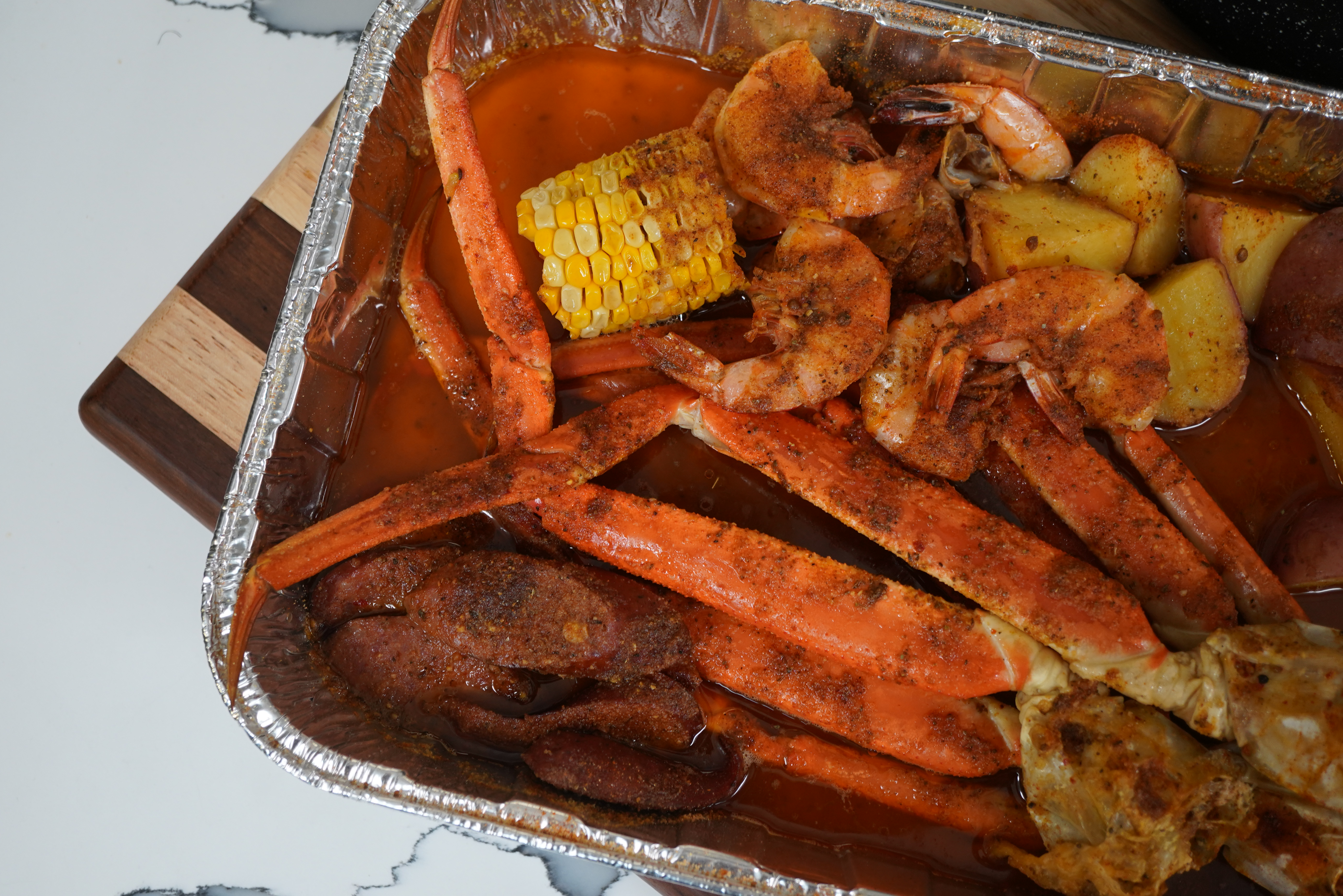 A tray of seasoned shrimp, corn, and roasted potatoes in a foil container.