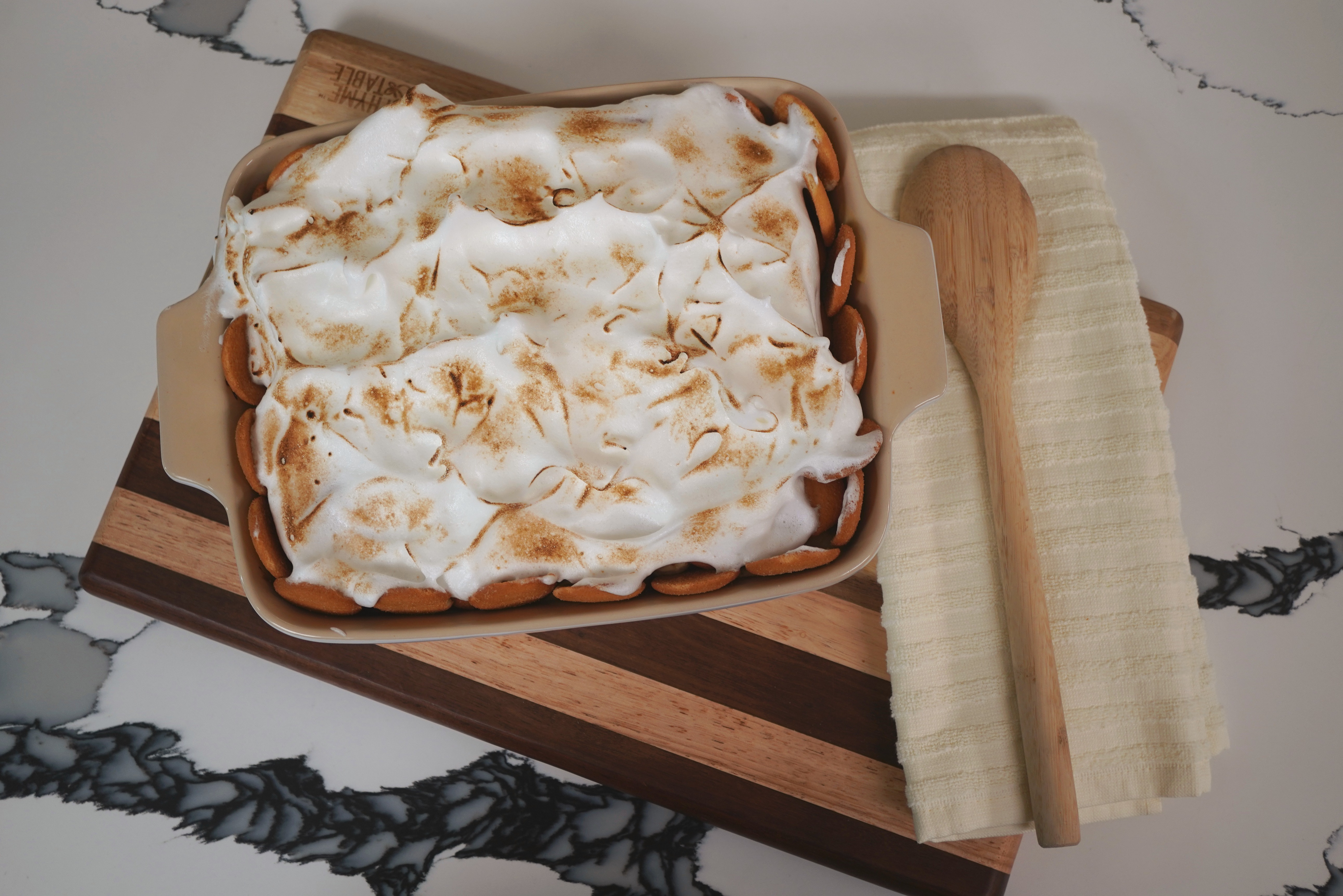 A wooden cutting board with a freshly baked sweet potato casserole topped with toasted marshmallow meringue, accompanied by a wooden spoon.
