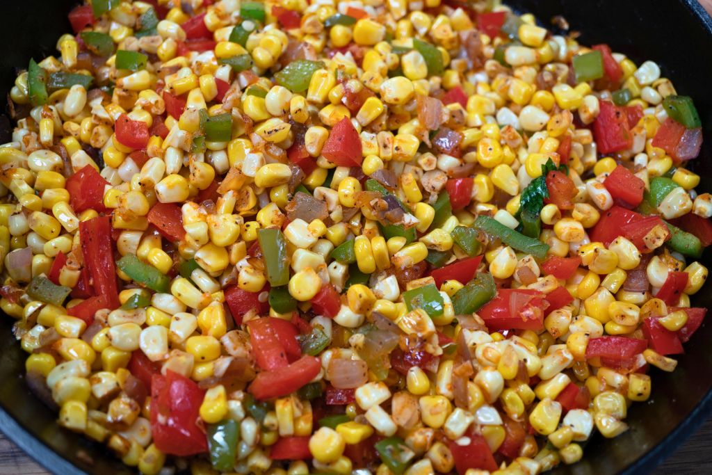 A close-up view of a pan filled with a colorful mix of sautéed corn kernels, red bell peppers, and other vegetables.
