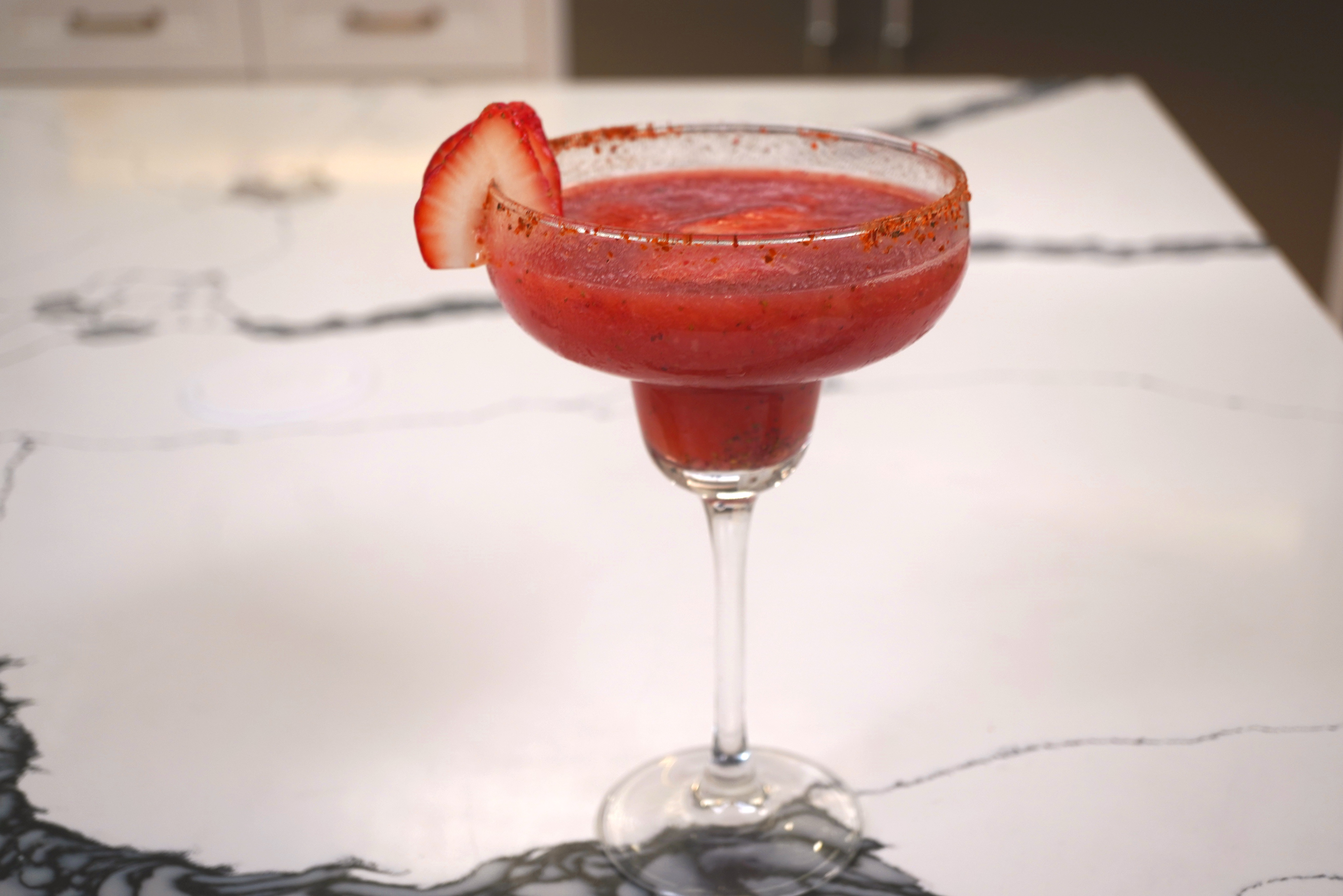 A red strawberry garnished margarita in a stemmed glass on a marble surface.