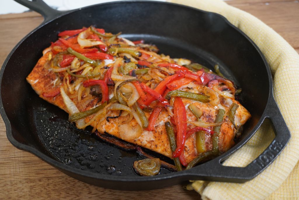A cast iron skillet filled with a sizzling dish of grilled salmon, bell peppers, and onions.