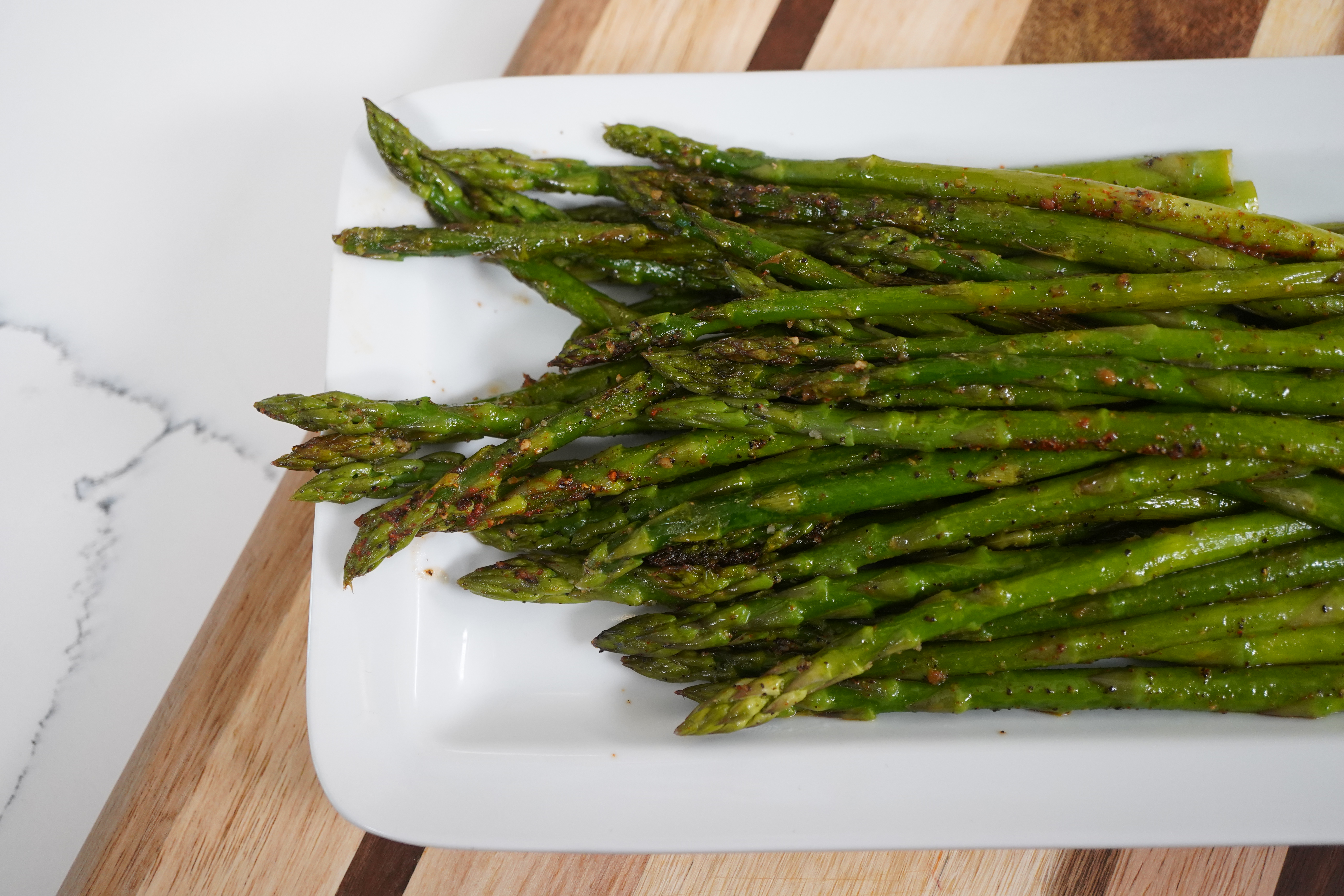Plate of roasted green asparagus spears with seasoning on a white plate and wooden surface.