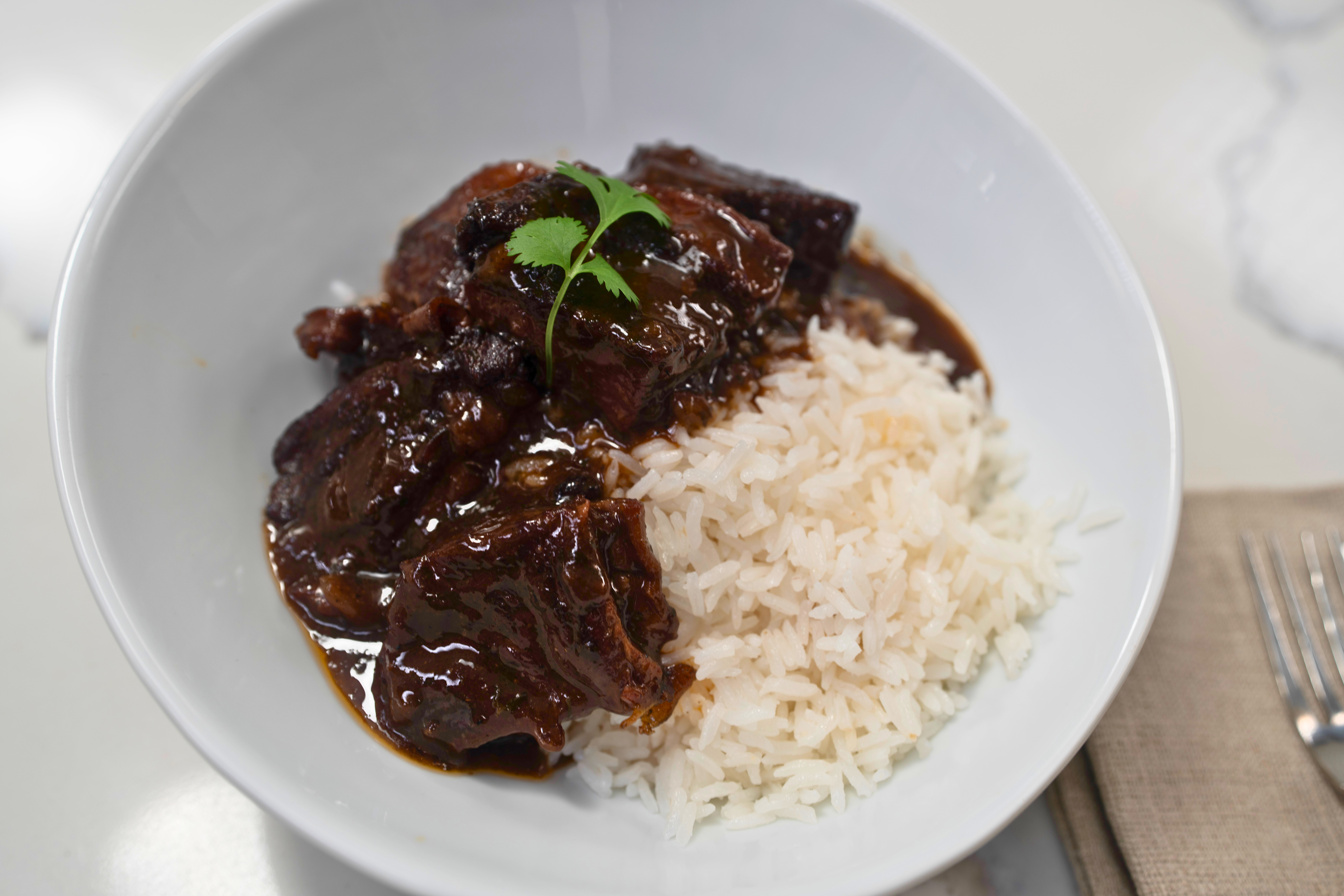 Braised beef short ribs served over white rice, garnished with a green herb sprig, on a white plate.