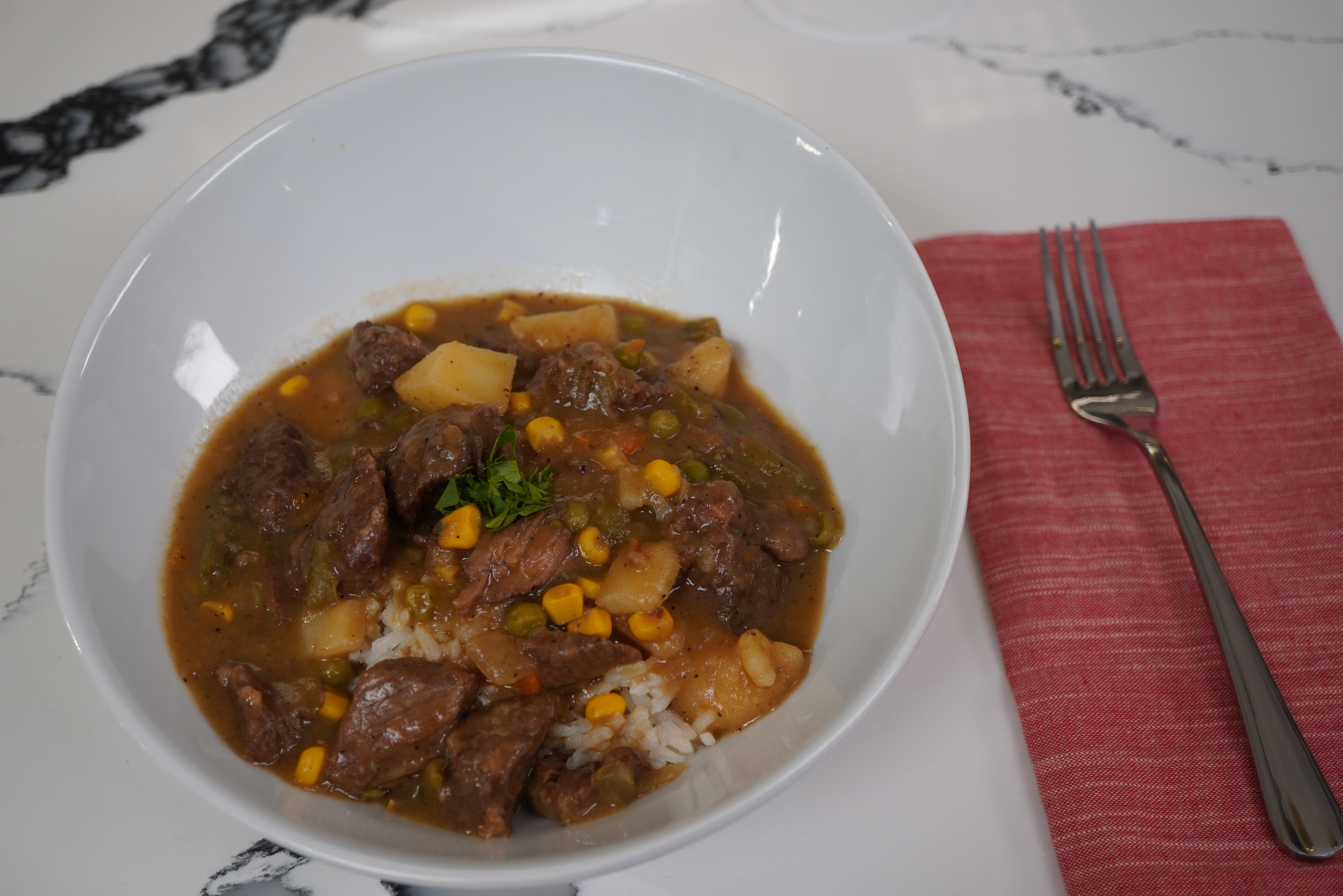 A bowl of beef stew with potatoes, corn, and herbs, served over rice on a marble background with a red cloth napkin and fork.