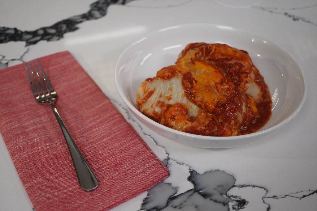 A white plate with a baked or fried chicken dish, possibly a chicken parmesan or chicken cacciatore, served with a red napkin and a fork.