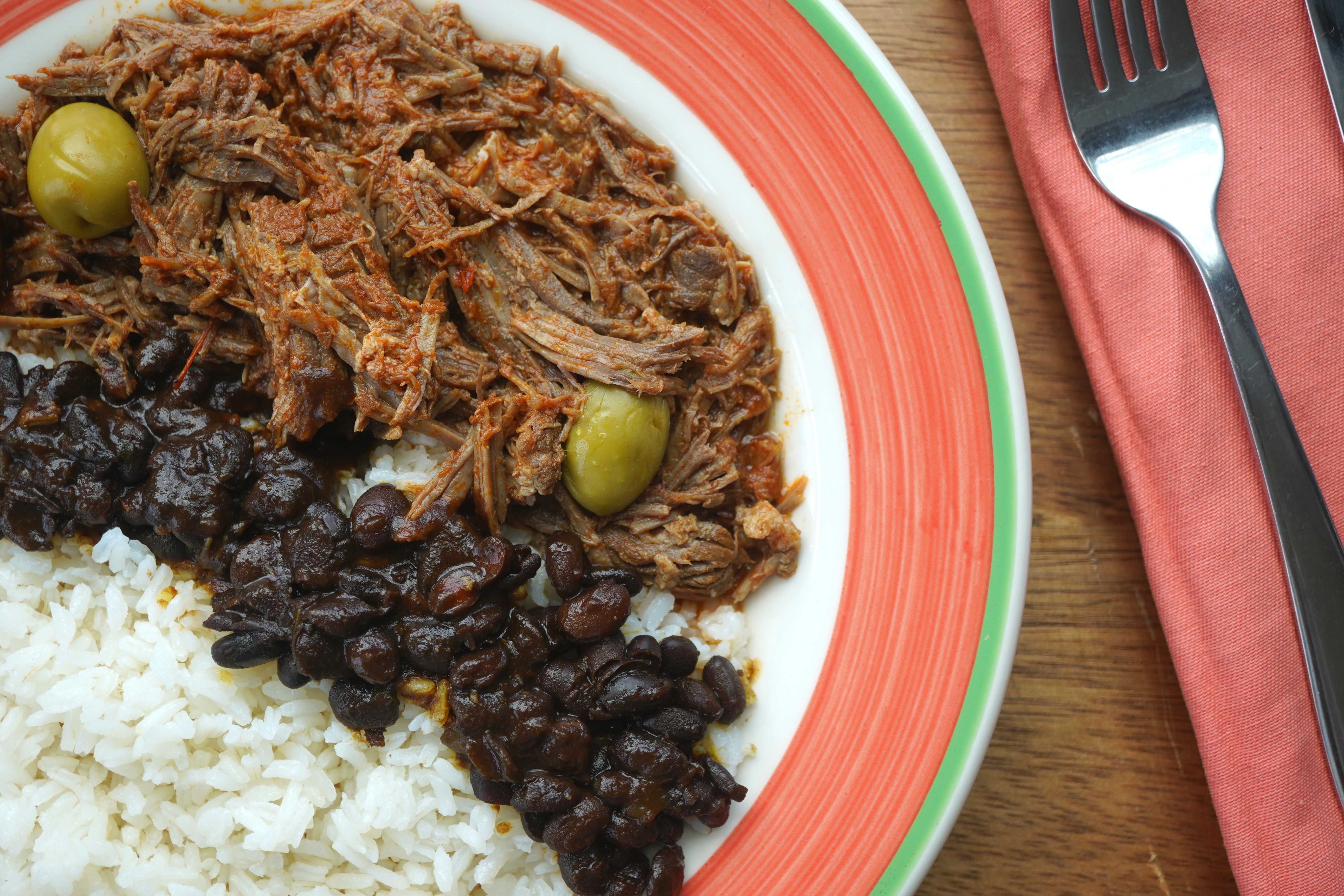 A plate of shredded meat, black beans, and white rice, garnished with green olives, on a red and green plate with a fork.