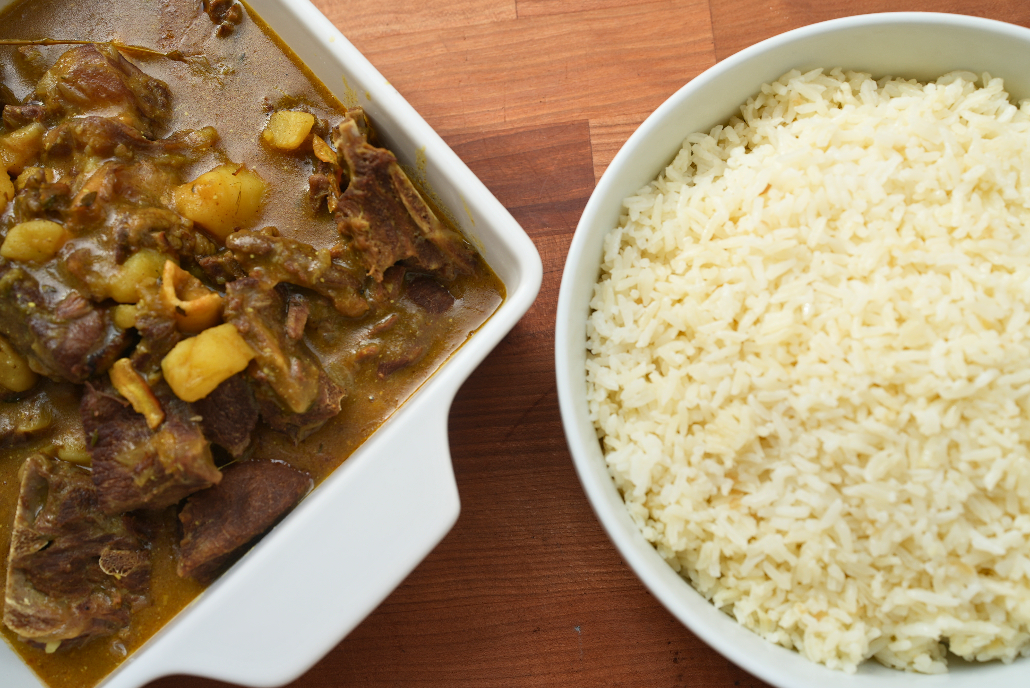 A white dish of beef stew with potatoes and a bowl of white rice on a wooden table.