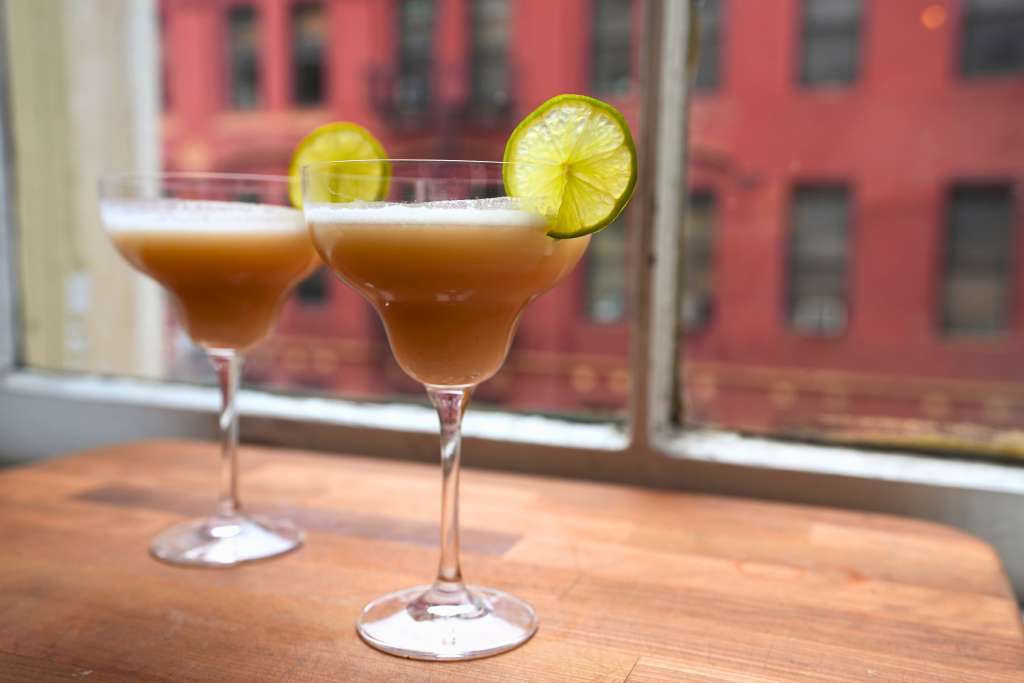 Two glasses of orange cocktail garnished with lime slices on a wooden table in front of a red brick building.